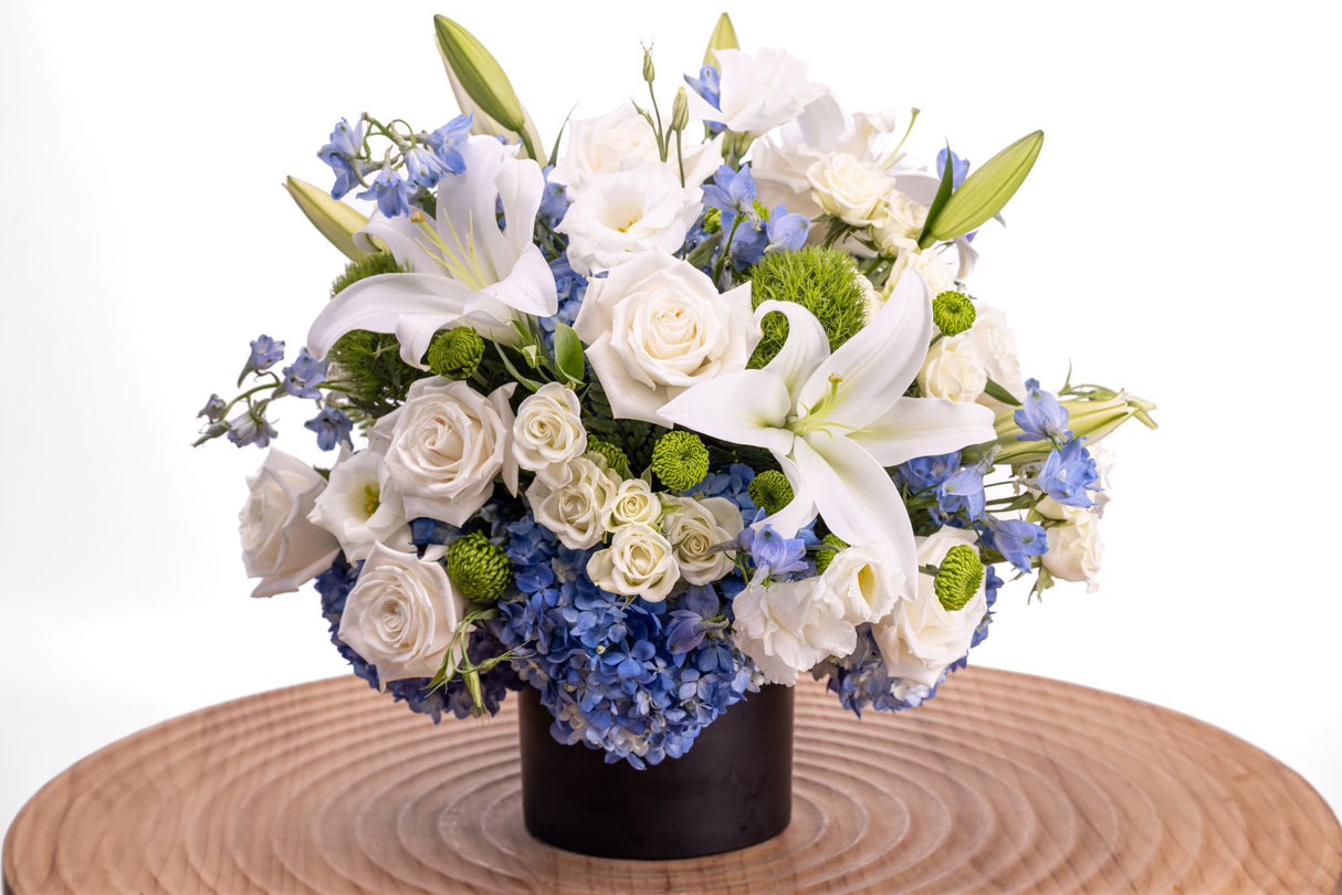 Blue Note features white roses, oriental lilies, lisianthus, and lush blue hydrangea accented with blue delphinium, green trick dianthus, button poms, and leatherleaf, arranged in a sleek black ceramic vase.