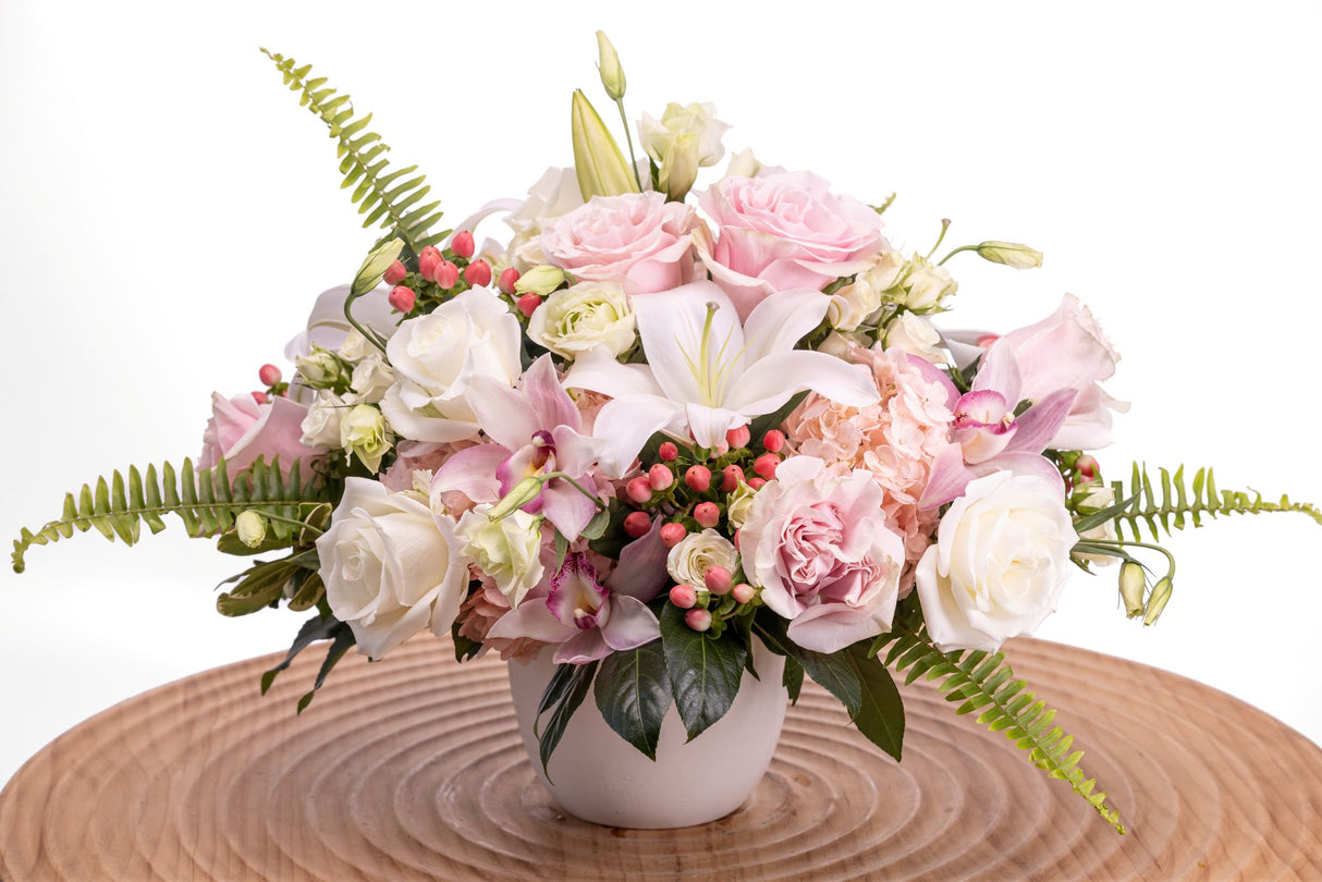 Elegance features white and pink roses, pink cymbidium orchids, white lilies, light pink hydrangea, lisianthus, hypericum, and lush greenery arranged in a white ceramic vase.