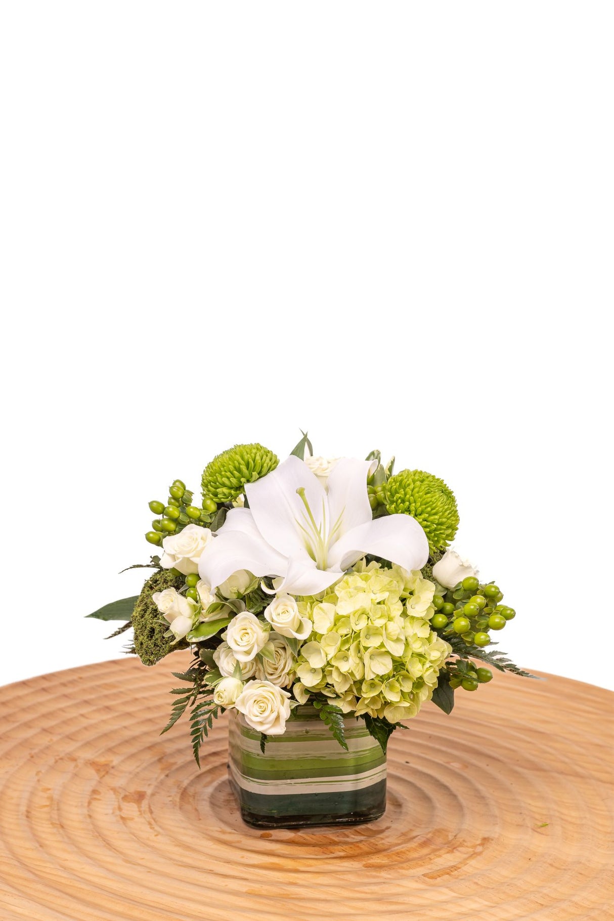 Emerald features white lilies with green hydrangea, pom cushions, hypericum berries, and lotus pods in a clear glass cube vase.