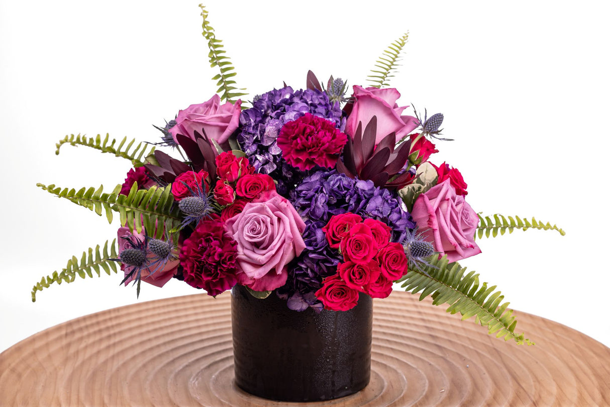 Royalty features lavender roses, hot pink spray roses, purple carnations, tinted hydrangea, thistle, safari sunset, and lush greenery arranged in a sleek black ceramic vase.