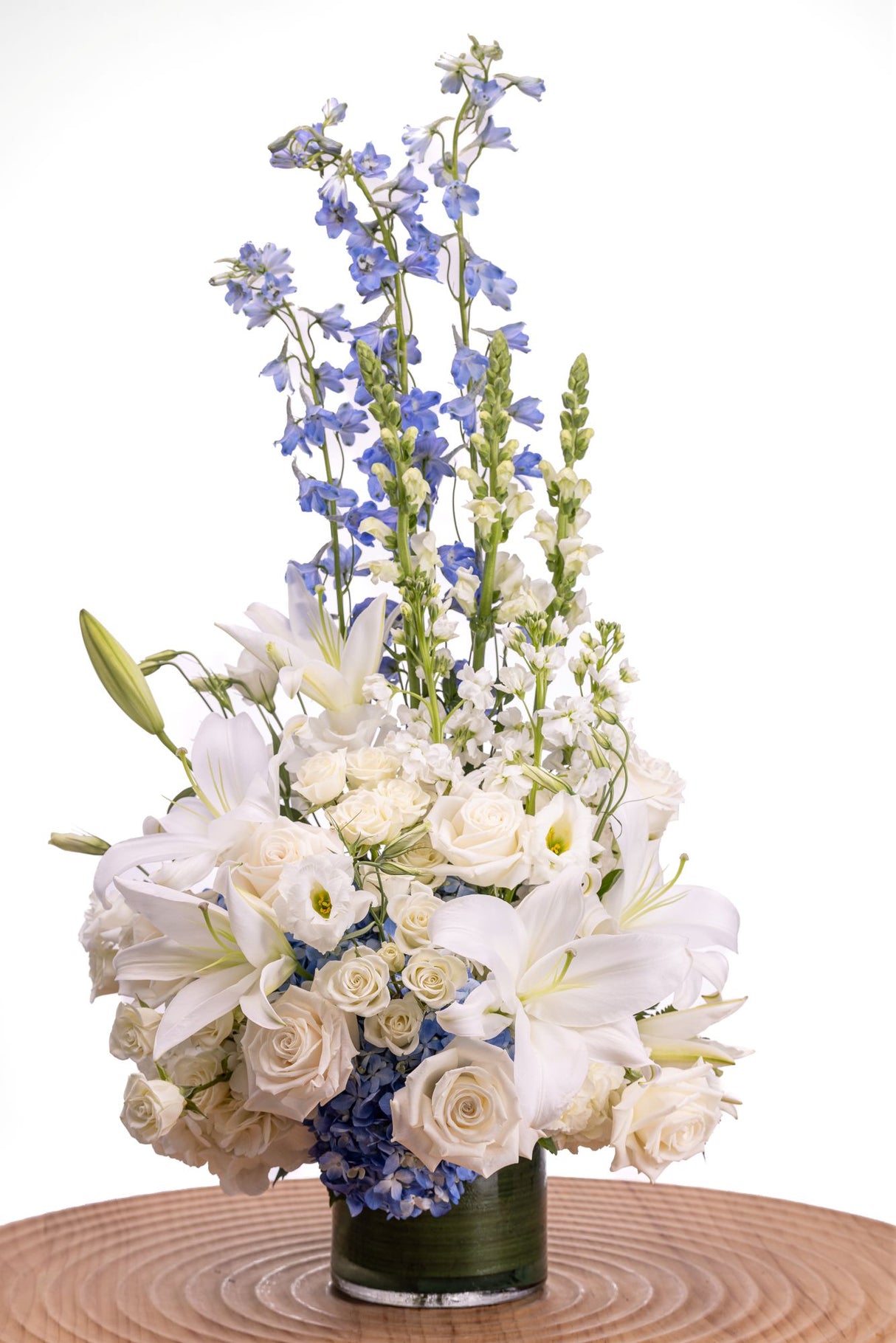 Skyfall features tall blue delphinium and white snapdragons arranged with white roses, lilies, lisianthus, hydrangea, and greenery in a clear glass cylinder vase.
