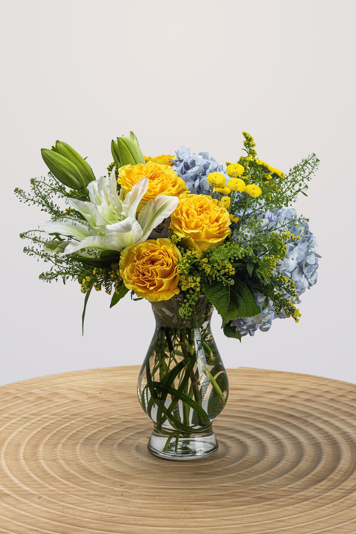 Arrangement featuring blue, yellow, white, and green hues of lilies, hydrangeas, and lush greenery.