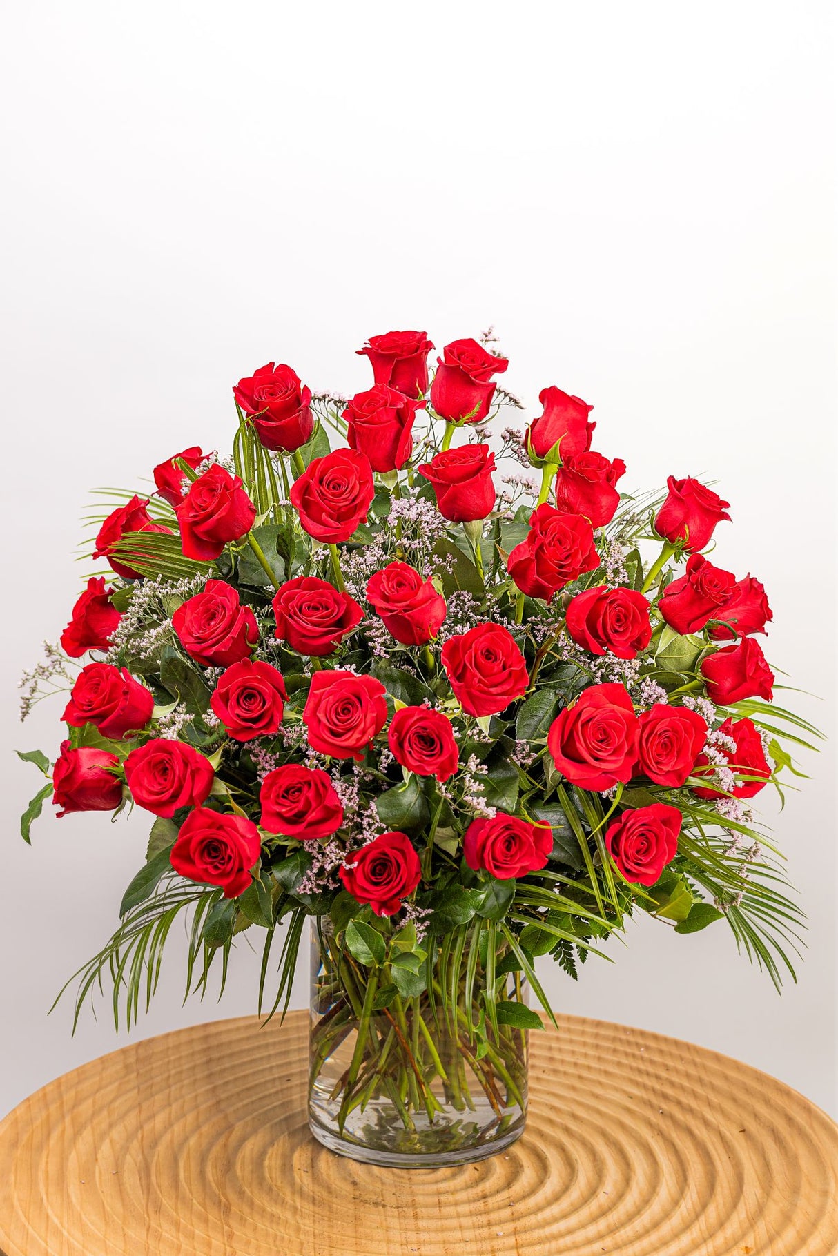Three Dozen Premium Red Roses features thirty-six long-stem red roses with soft pink limonium, robellini, leatherleaf, and salal, displayed in a cylinder glass vase.
