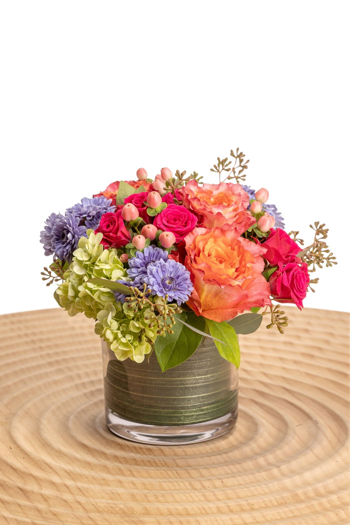 Viva features Free Spirit roses, hot pink spray roses, green hydrangea, peach hypericum berries, blue cushion poms, seeded eucalyptus and salal in a glass cylinder vase.