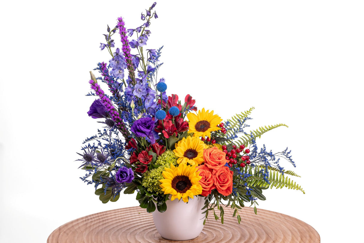 Vivid Blooms features sunflowers, orange roses, purple lisianthus, red alstroemeria, tinted craspedia, blue delphinium, and green hydrangea arranged in a white ceramic vase.