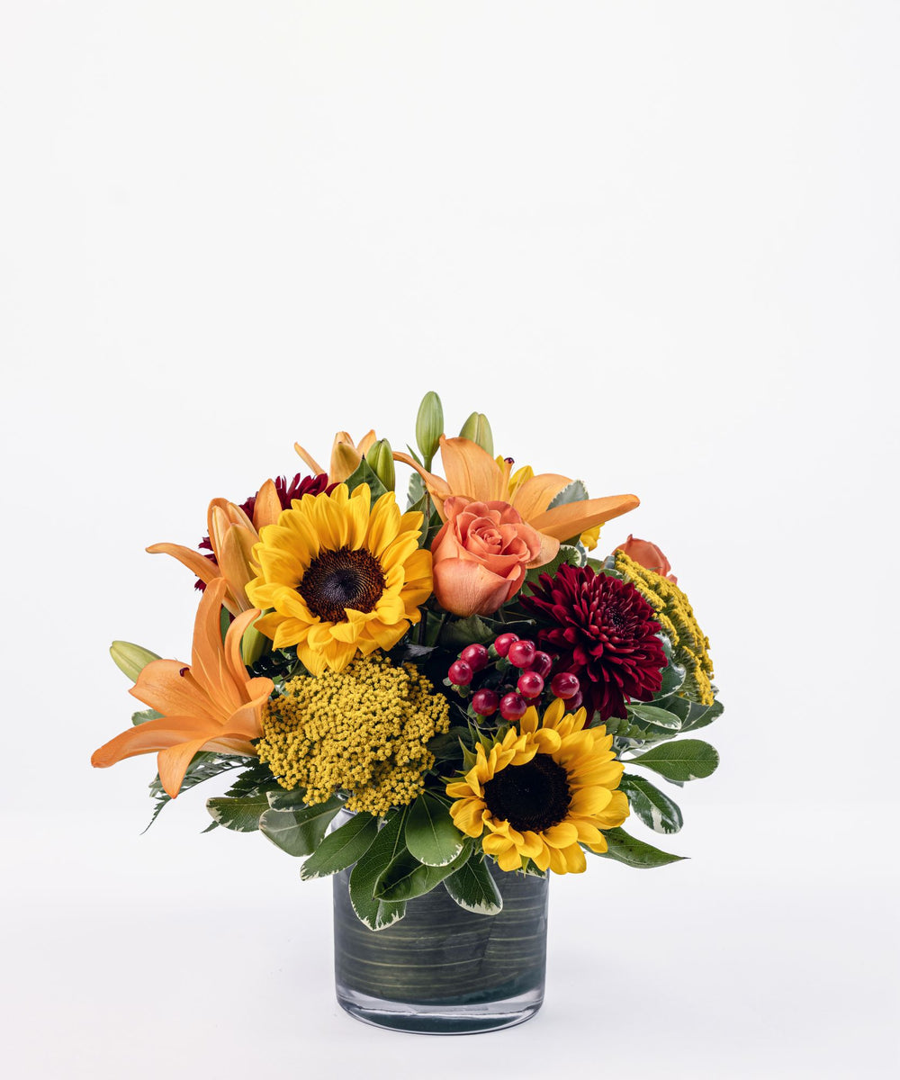 Autumn floral arrangement with sunflowers, orange roses, orange lilies, burgundy cremons, and red hypericum in a clear cylinder vase
