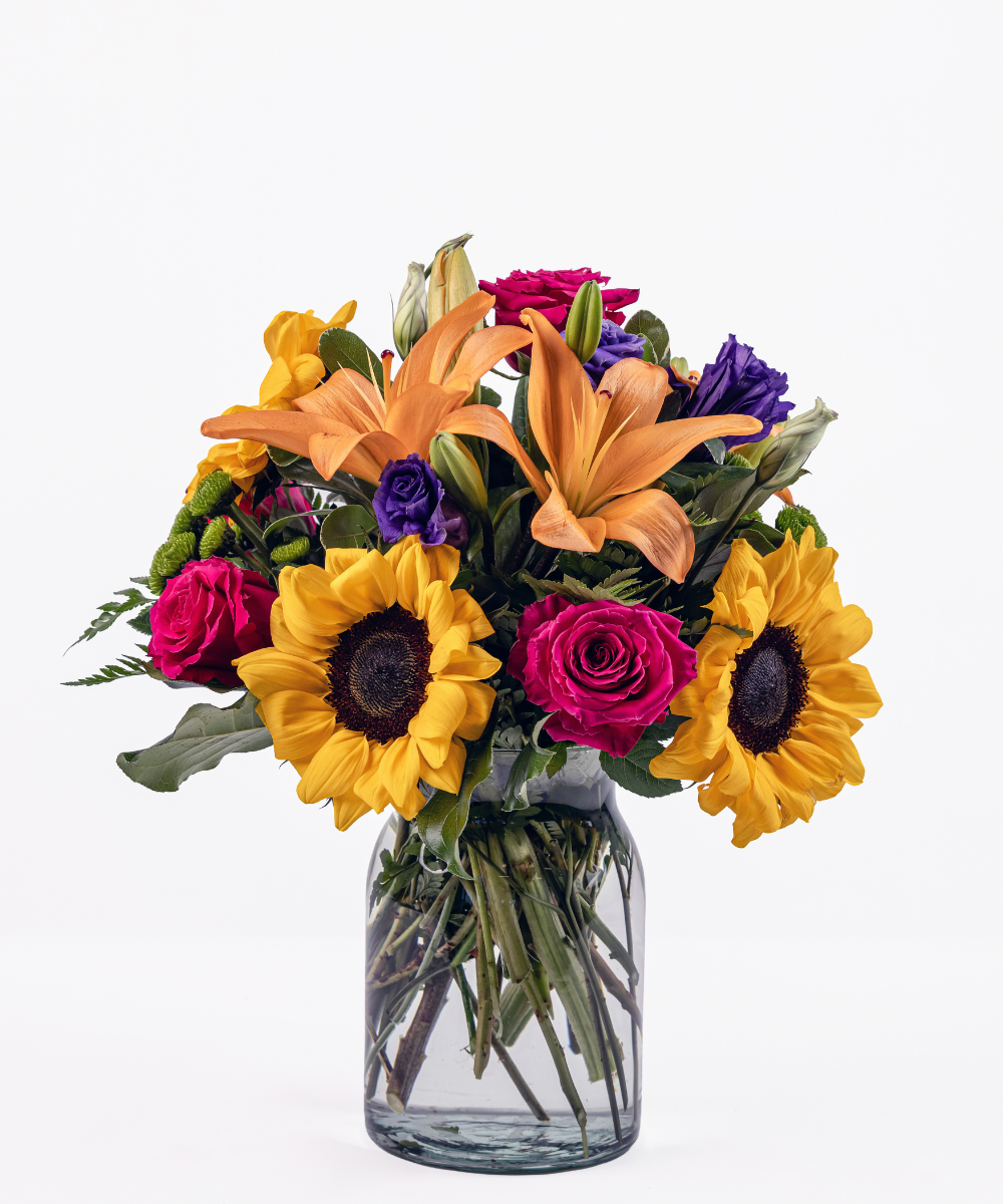 Cheerful mixed floral arrangement with sunflowers, orange lilies, hot pink roses, and purple lisianthus in a milk jug vase
