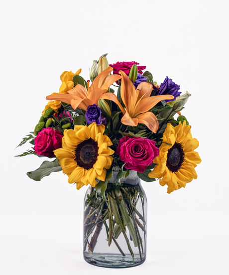 Cheerful mixed floral arrangement with sunflowers, orange lilies, hot pink roses, and purple lisianthus in a milk jug vase