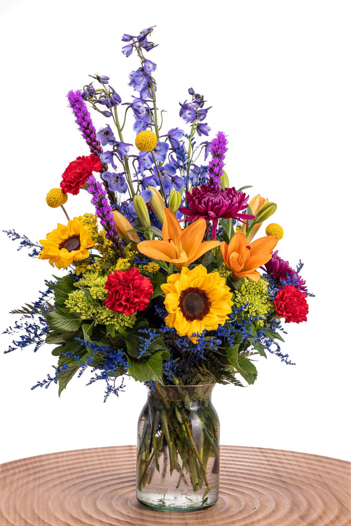 Bright Spirit features sunflowers, orange lilies, red carnations, purple cremons, blue delphinium, craspedia, liatris, solidago, and green hydrangea arranged in a glass vase.