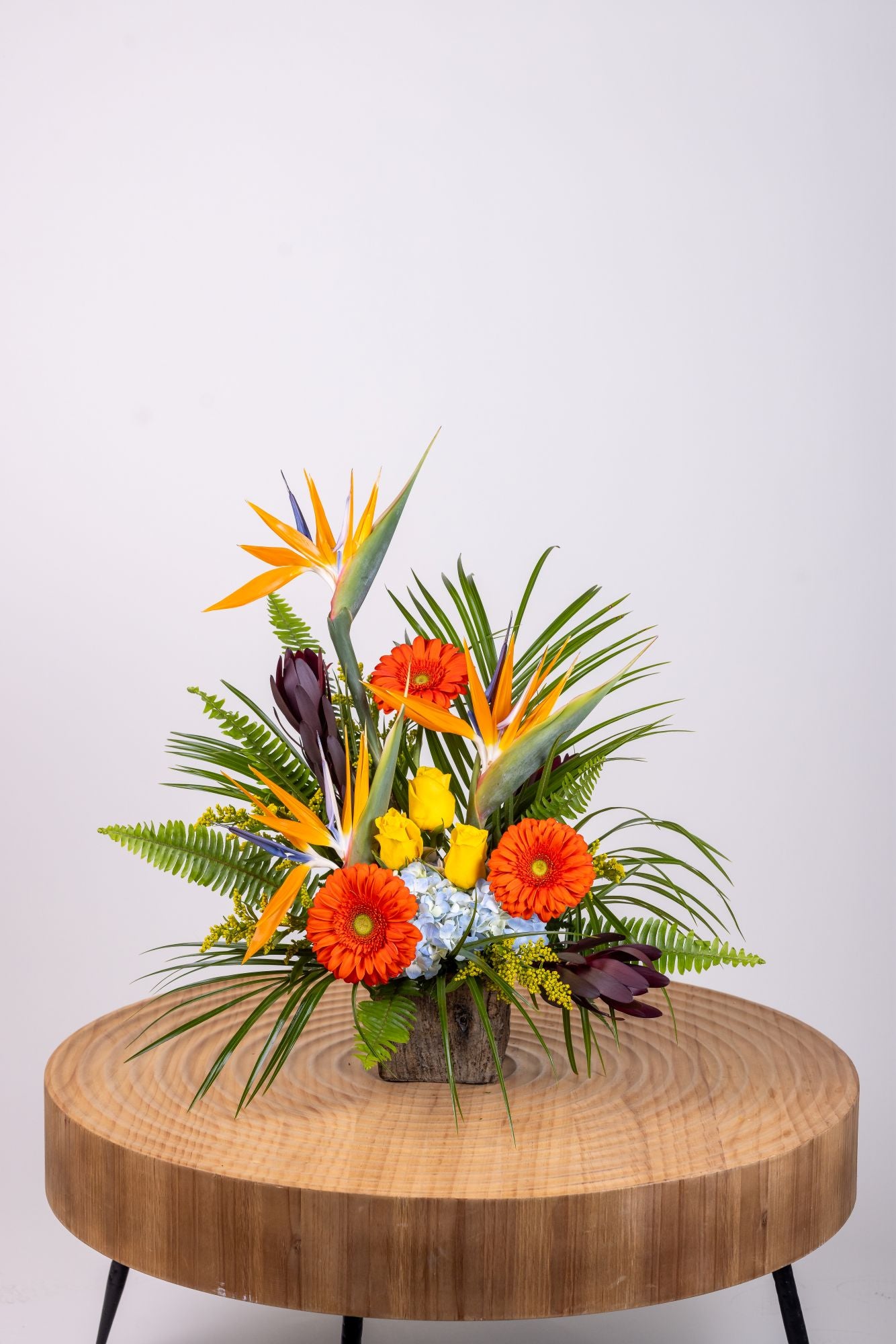 Tropical arrangement with birds of paradise, blue hydrangeas, yellow roses, and orange gerbera daisies in a wooden box