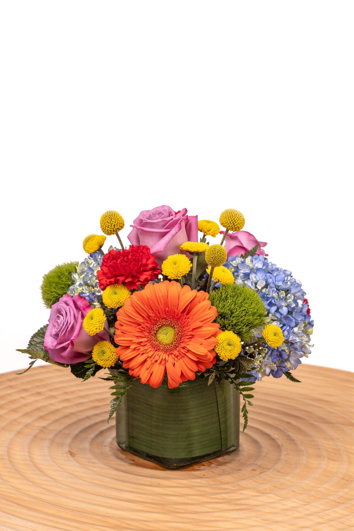 Chasing Rainbows features orange gerbera daisies, lavender roses, red carnations, blue hydrangea, yellow craspedia, and green trick arranged in a glass cube vase.
