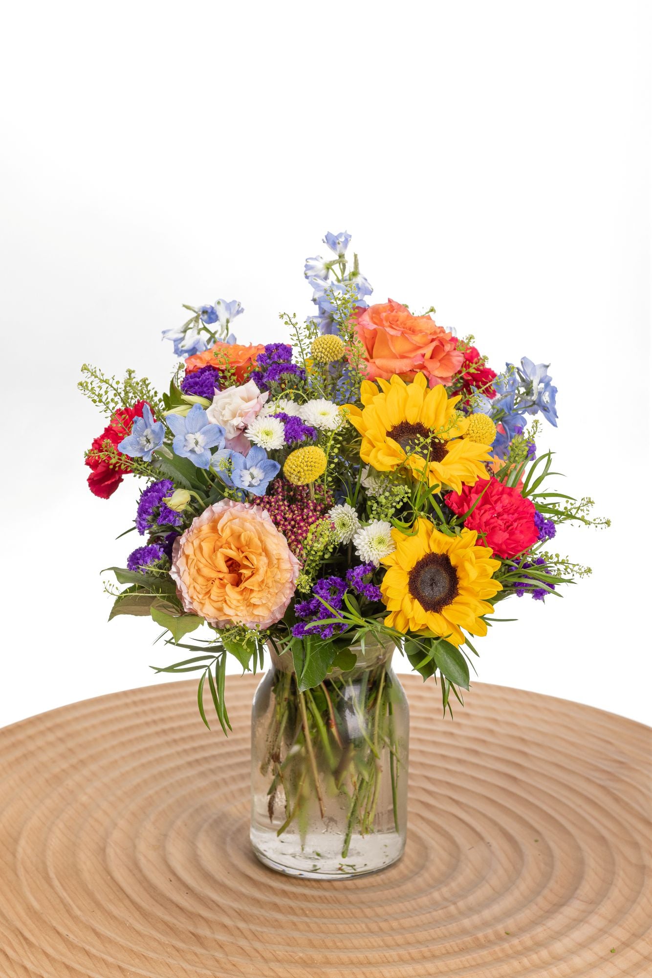 Chromatic features sunflowers, Free Spirit roses, pink carnations and blue delphinium in a glass milk jug vase.
