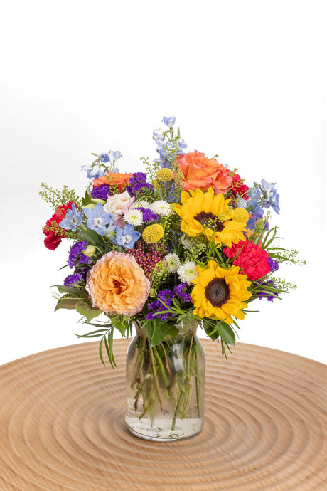 Chromatic features sunflowers, Free Spirit roses, pink carnations and blue delphinium in a glass milk jug vase.