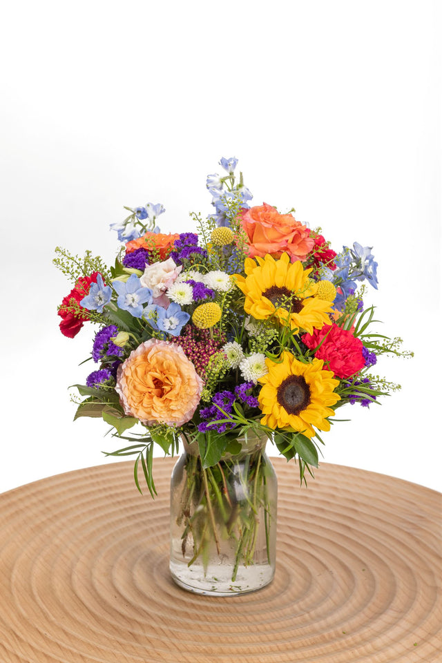 Chromatic features sunflowers, Free Spirit roses, pink carnations and blue delphinium in a glass milk jug vase.