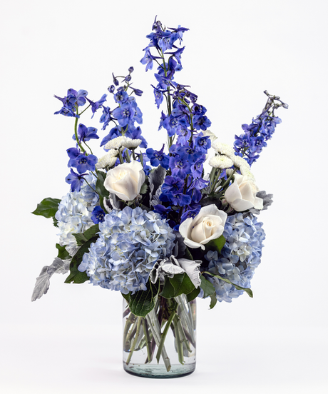 Blue and white floral arrangement with delphinium, hydrangea, white roses, and button poms in a milk jug vase