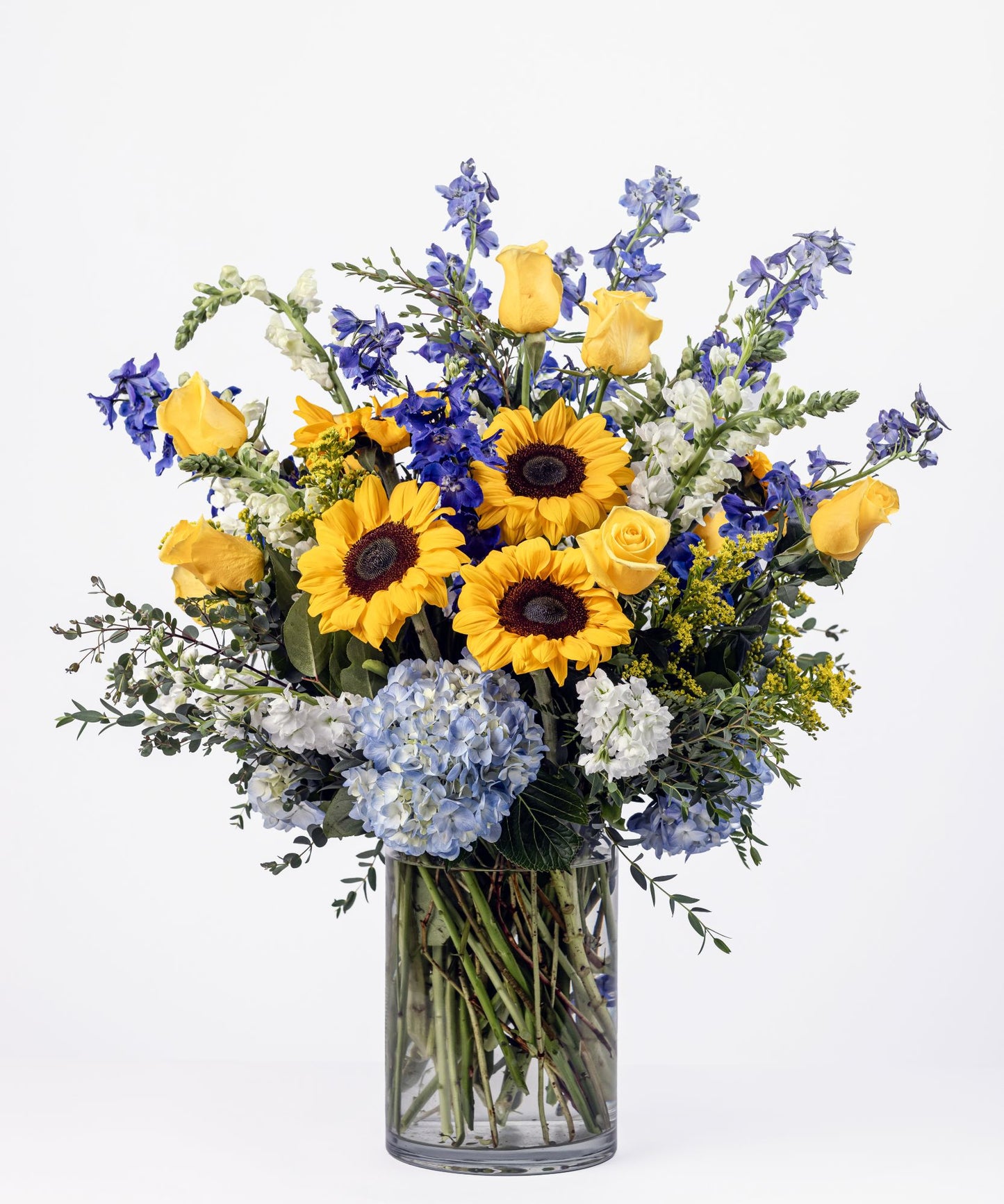 Bright floral arrangement with sunflowers, blue hydrangea, dark blue delphinium, yellow roses, white snapdragons, and white stock in a clear glass vase