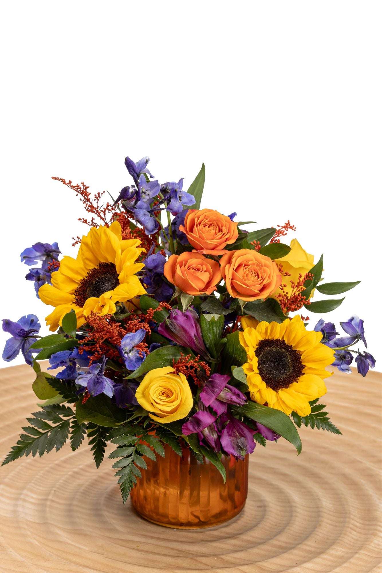 Color Fall features golden sunflowers, orange spray roses, yellow roses, purple alstroemeria, and blue delphinium, accented with tinted solidago and greenery, arranged in a warm marigold ribbed glass jar.