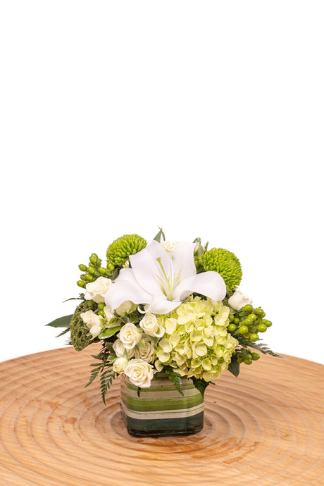 Emerald features white lilies with green hydrangea, pom cushions, hypericum berries, and lotus pods in a clear glass cube vase.