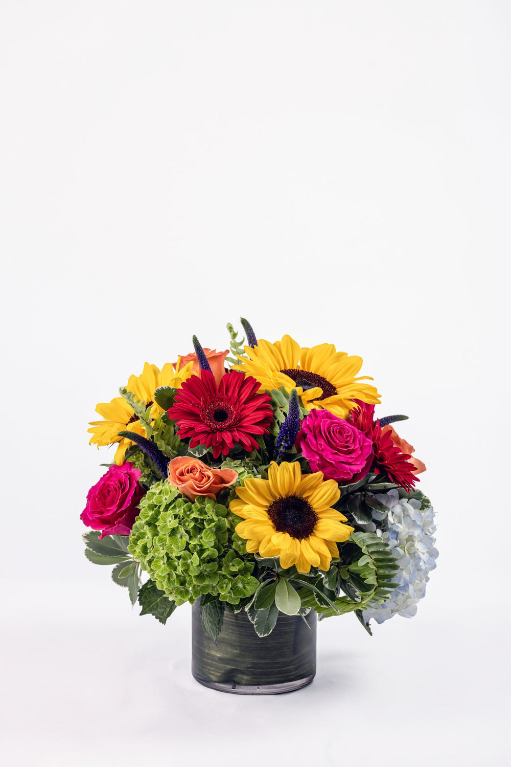 Vibrant fall-inspired floral arrangement with hydrangea, sunflowers, roses, mini gerbera daisies, and purple veronica in a clear cylinder vase