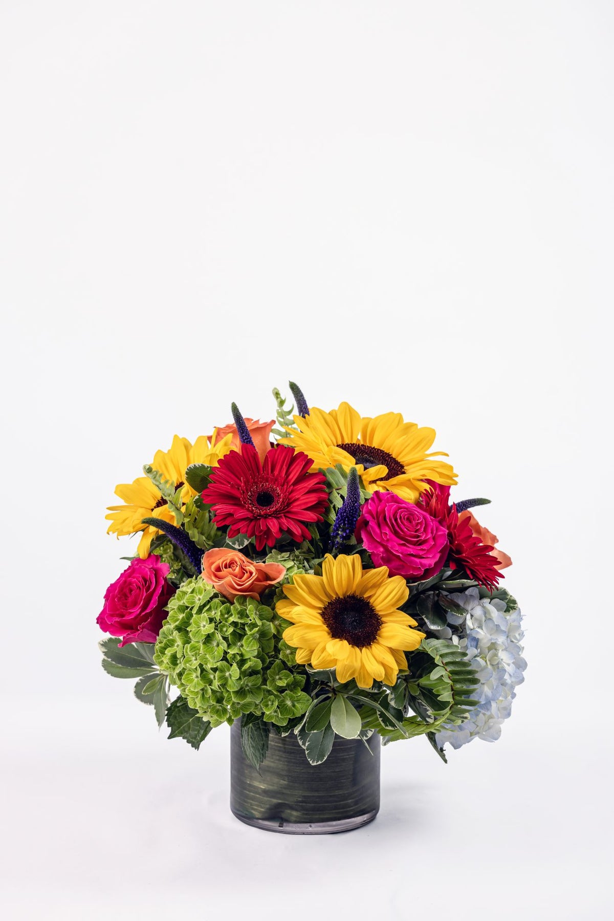 Vibrant fall-inspired floral arrangement with hydrangea, sunflowers, roses, mini gerbera daisies, and purple veronica in a clear cylinder vase