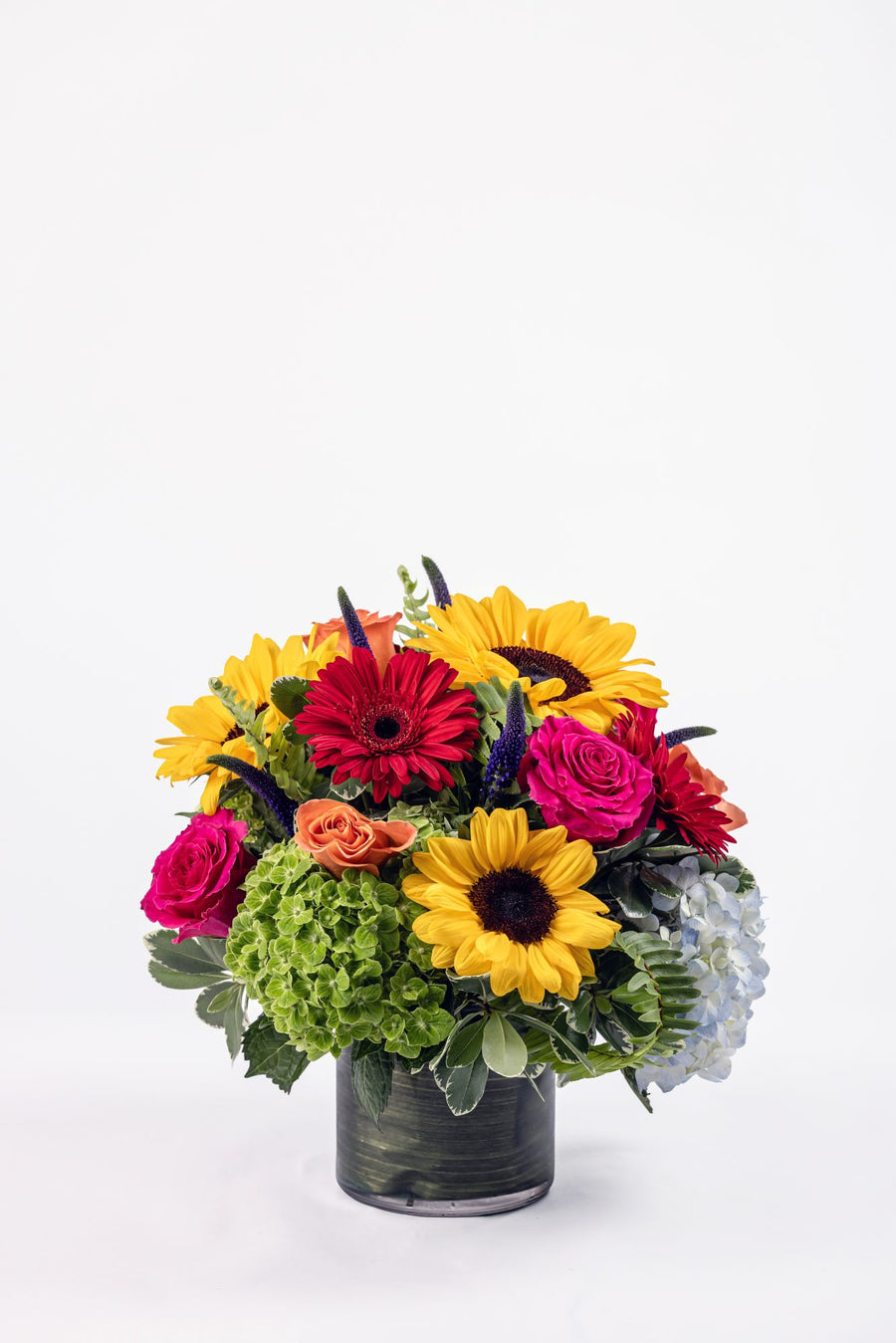Vibrant fall-inspired floral arrangement with hydrangea, sunflowers, roses, mini gerbera daisies, and purple veronica in a clear cylinder vase