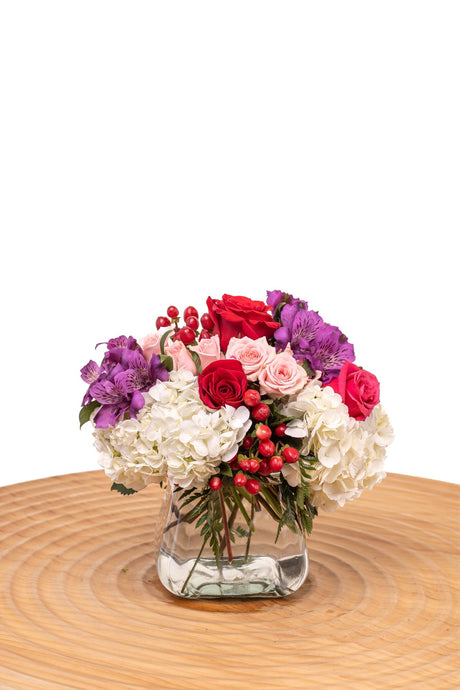 First Kiss features red and pink roses, spray roses, white hydrangea, and purple alstroemeria with red hypericum berries in a glass cube vase.