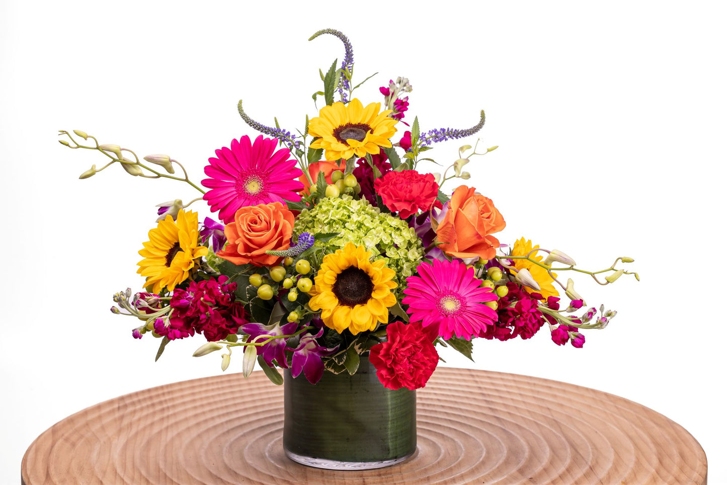 Flair features sunflowers, hot pink gerbera daisies, orange roses, pink dendrobium orchids, carnations, stock, hydrangea, veronica, and hypericum arranged in a glass cylinder vase wrapped with ribbon leaf.
