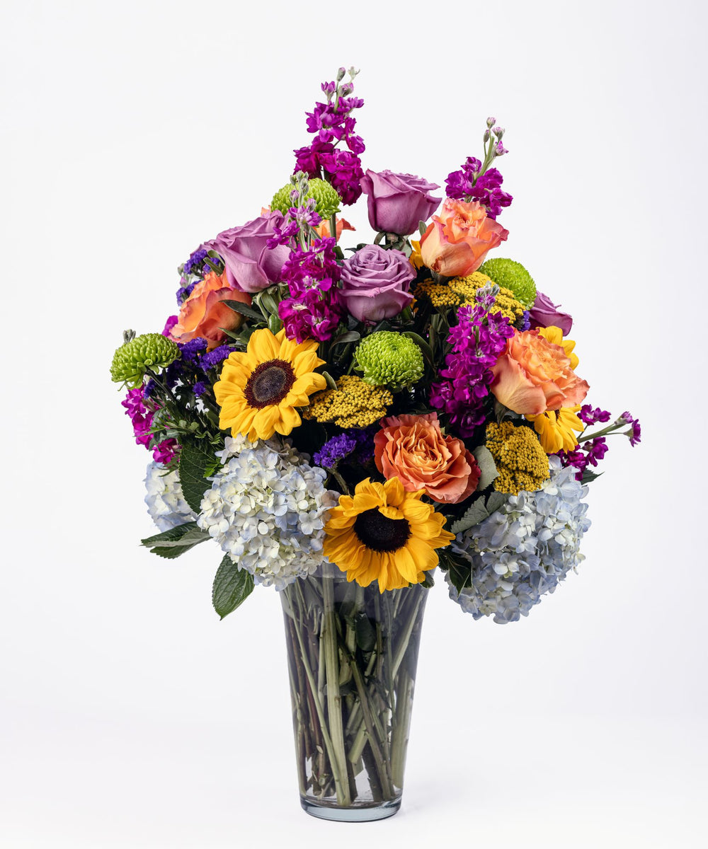 Colorful garden-style floral arrangement with sunflowers, blue hydrangea, lavender roses, Free Spirit roses, purple stock, and green cremons in a Riviera vase