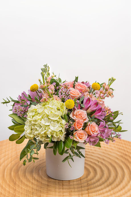 Garden Party features white hydrangea, peach spray roses, purple alstroemeria, peach stock, yellow craspedia, pink waxflower, and fresh greenery arranged in a white ceramic vase.
