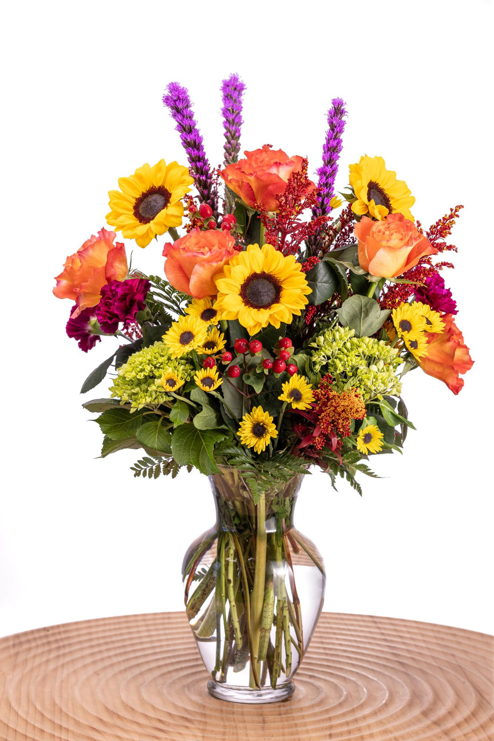 In Fall Bloom features golden sunflowers, orange Free Spirit roses, purple carnations, liatris, and Viking poms, accented with red hypericum, solidago, mini green hydrangea, and lush greenery in a glass vase.