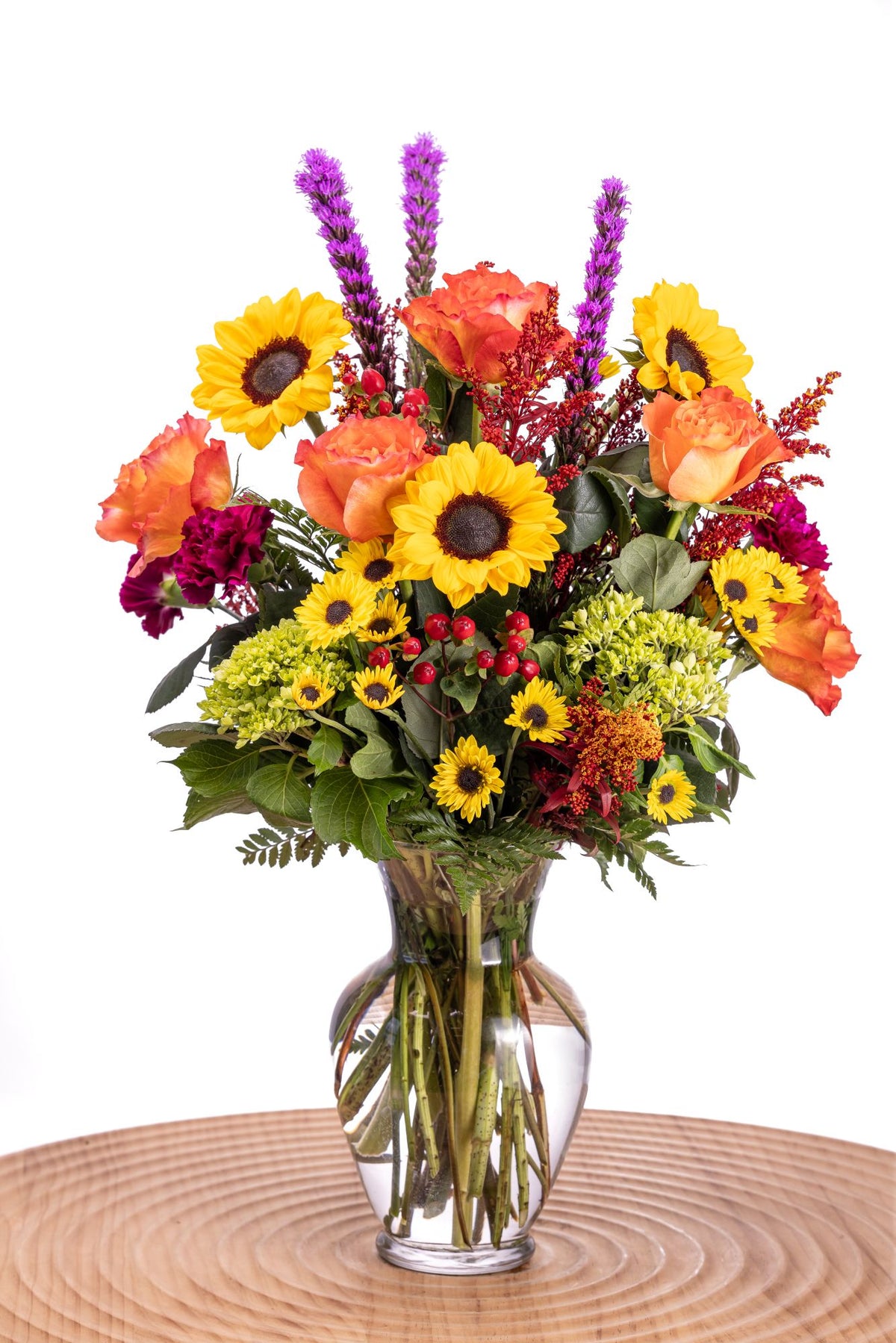 In Fall Bloom features golden sunflowers, orange Free Spirit roses, purple carnations, liatris, and Viking poms, accented with red hypericum, solidago, mini green hydrangea, and lush greenery in a glass vase.