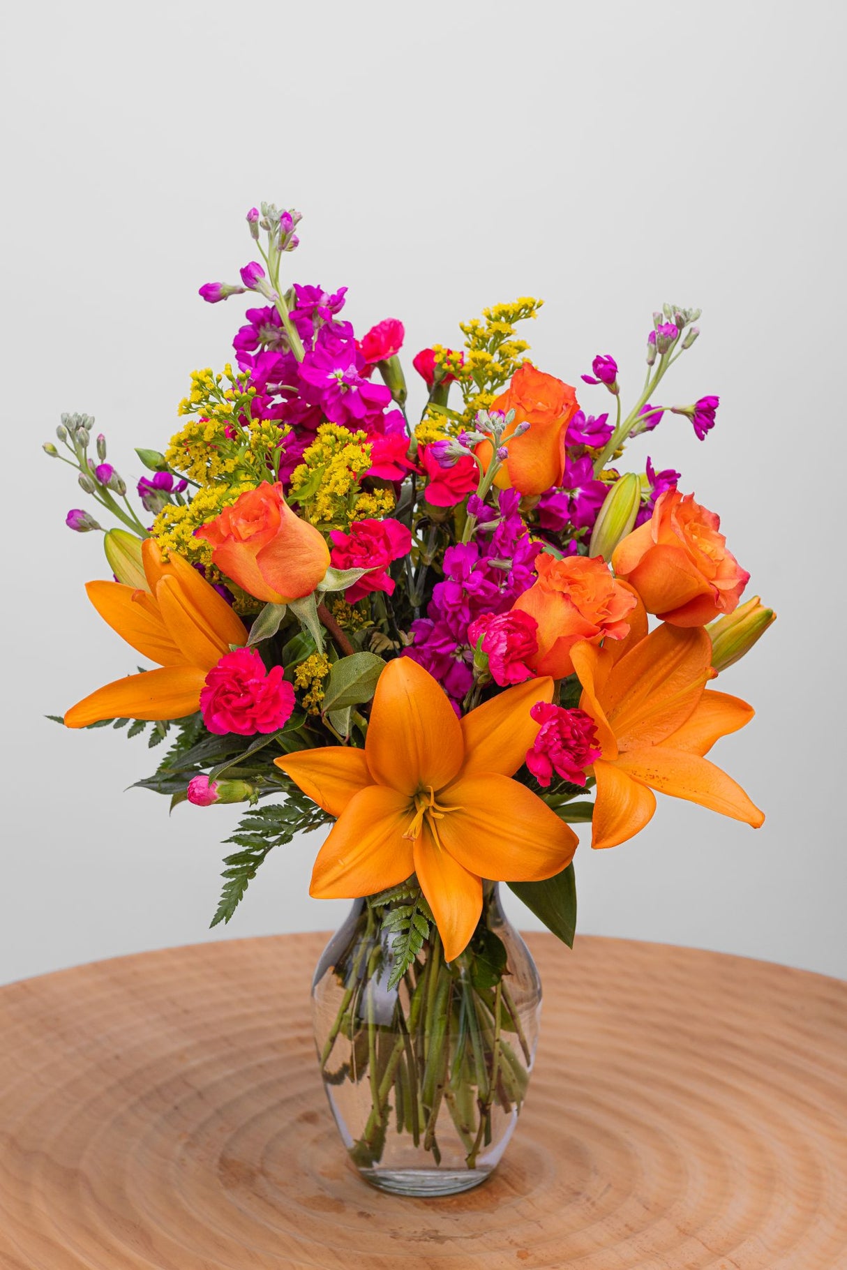 Just Happy Bouquet features orange lilies and roses, fuchsia stock and mini carnations, solidago, and lush greenery arranged in a glass rose vase.
