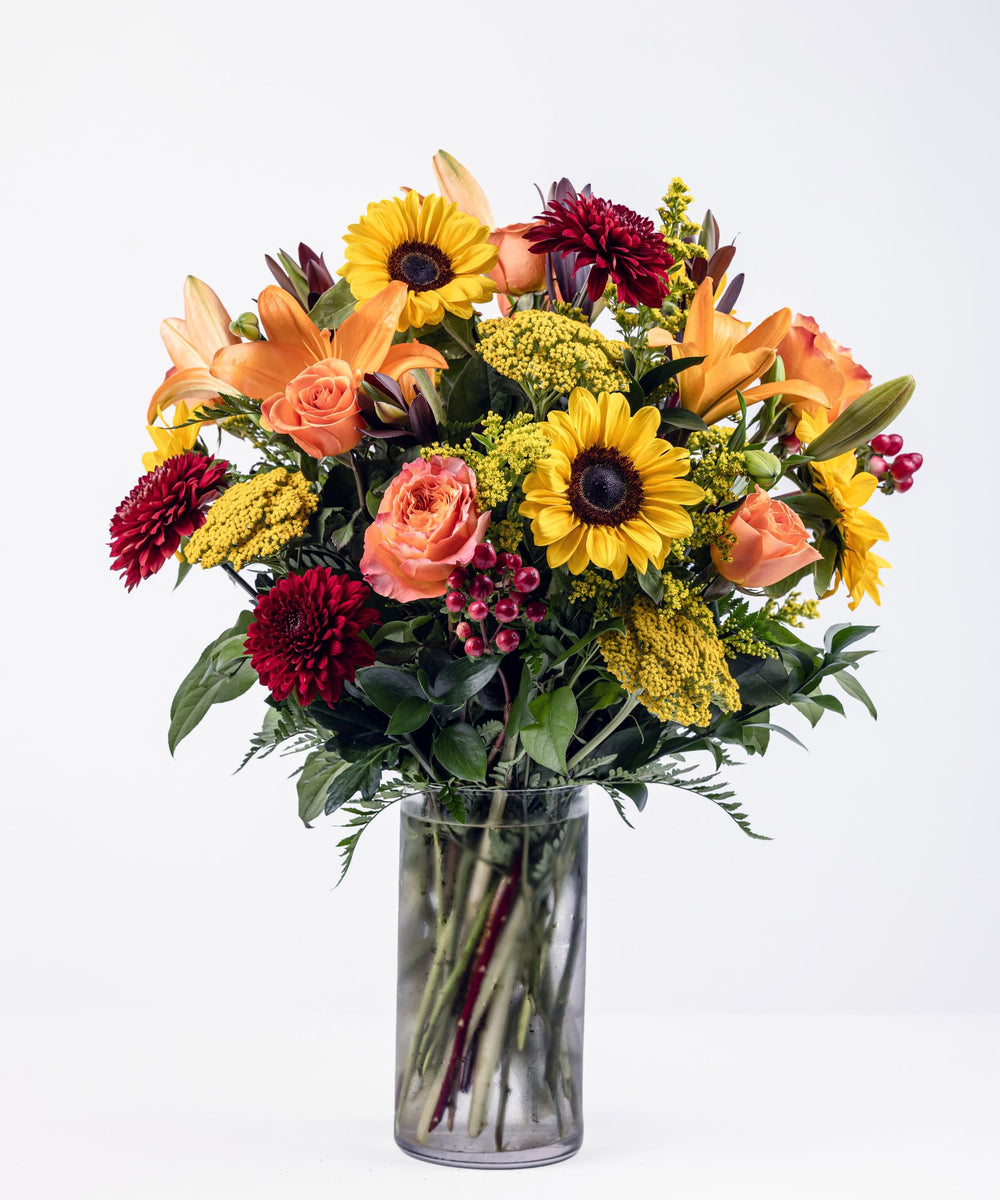 Autumn floral arrangement with sunflowers, orange lilies, Free Spirit roses, burgundy cremons, and red hypericum in a clear glass vase