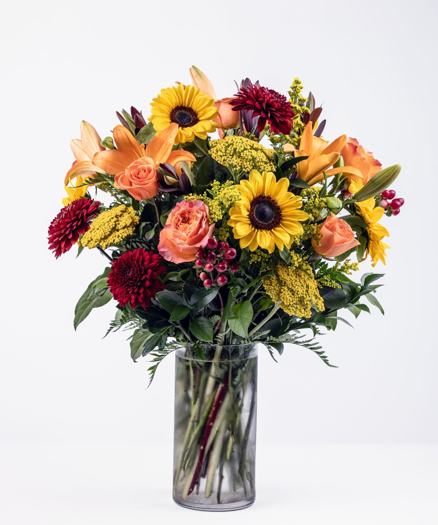 Autumn floral arrangement with sunflowers, orange lilies, Free Spirit roses, burgundy cremons, and red hypericum in a clear glass vase