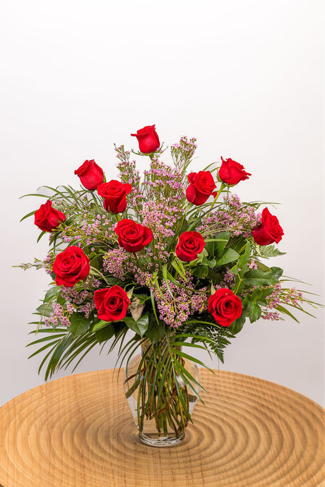 An exquisite flower arrangement featuring 12 long-stemmed red roses, small blooms of high quality filler flowers, and fresh greens in a tall vase.