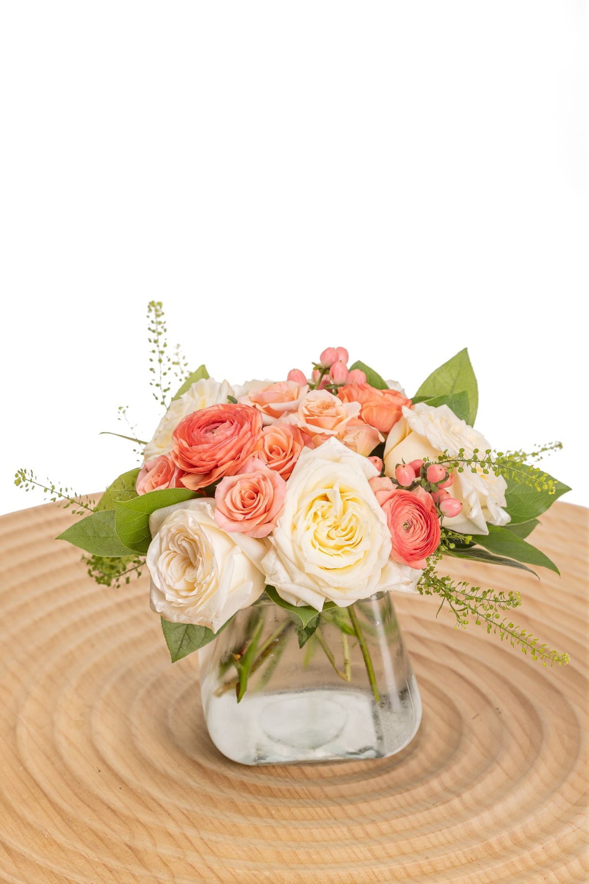 Peaches and Cream features cream and peach roses, peach ranunculus, blush spray roses, pink hypericum berries, pennycress, and fresh greenery arranged in a clear posie square vase.