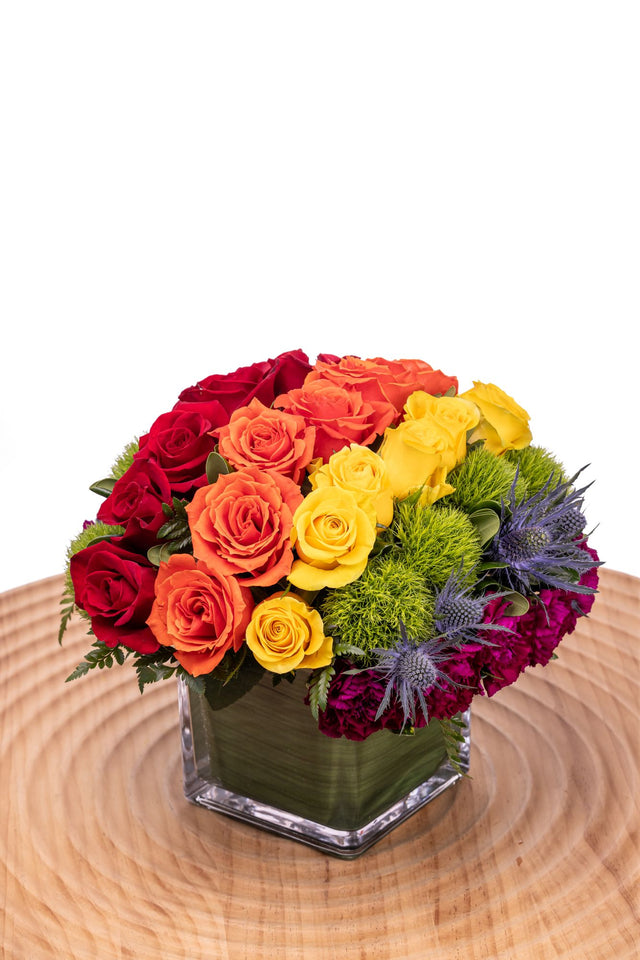 Chasing Rainbows features orange gerbera daisies, lavender roses, red carnations, blue hydrangea, yellow craspedia, and green trick arranged in a glass cube vase.
