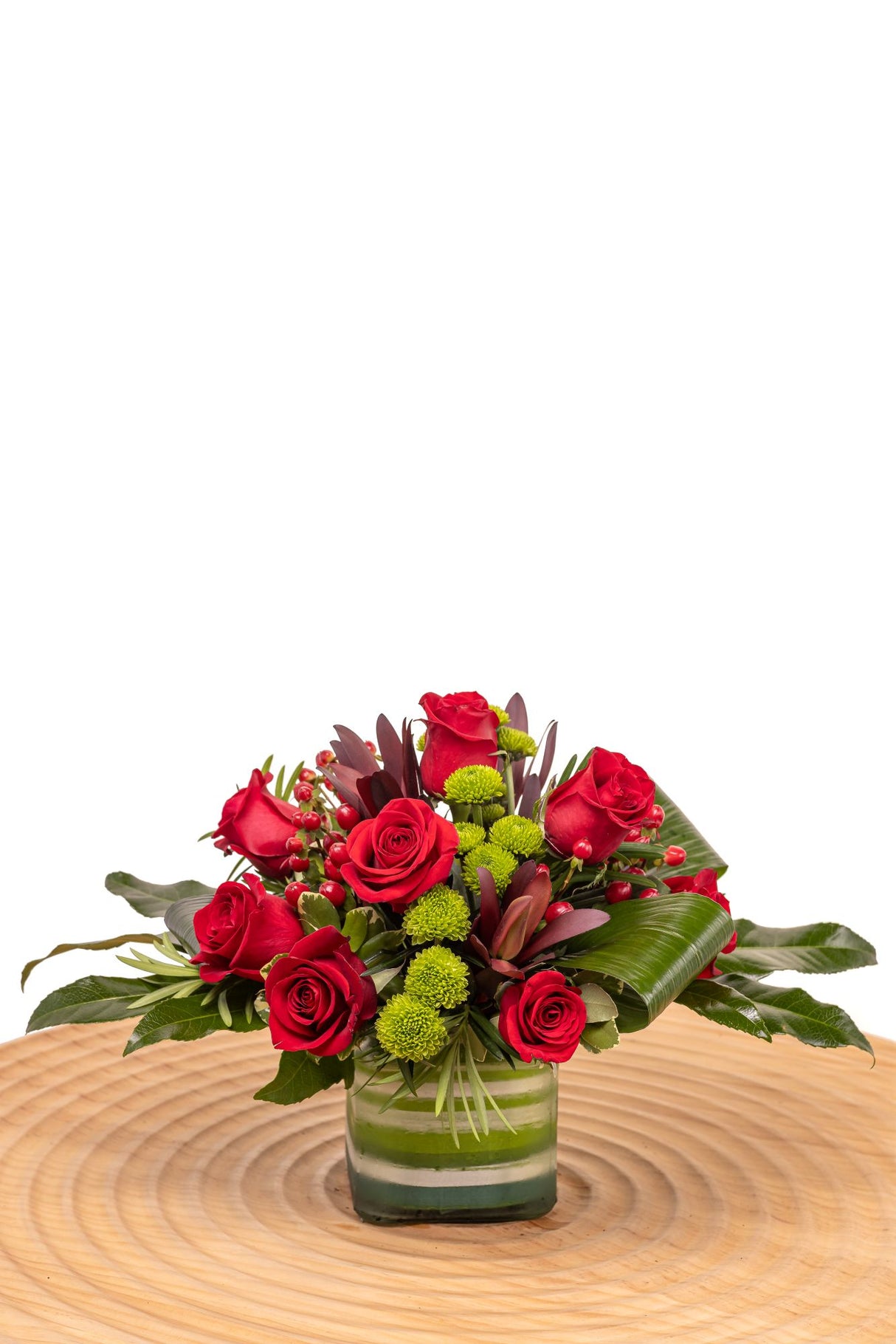 Ruby features red roses, safari sunset, green button poms, and assorted foliage arranged in a glass cube vase lined with ribbon leaf.