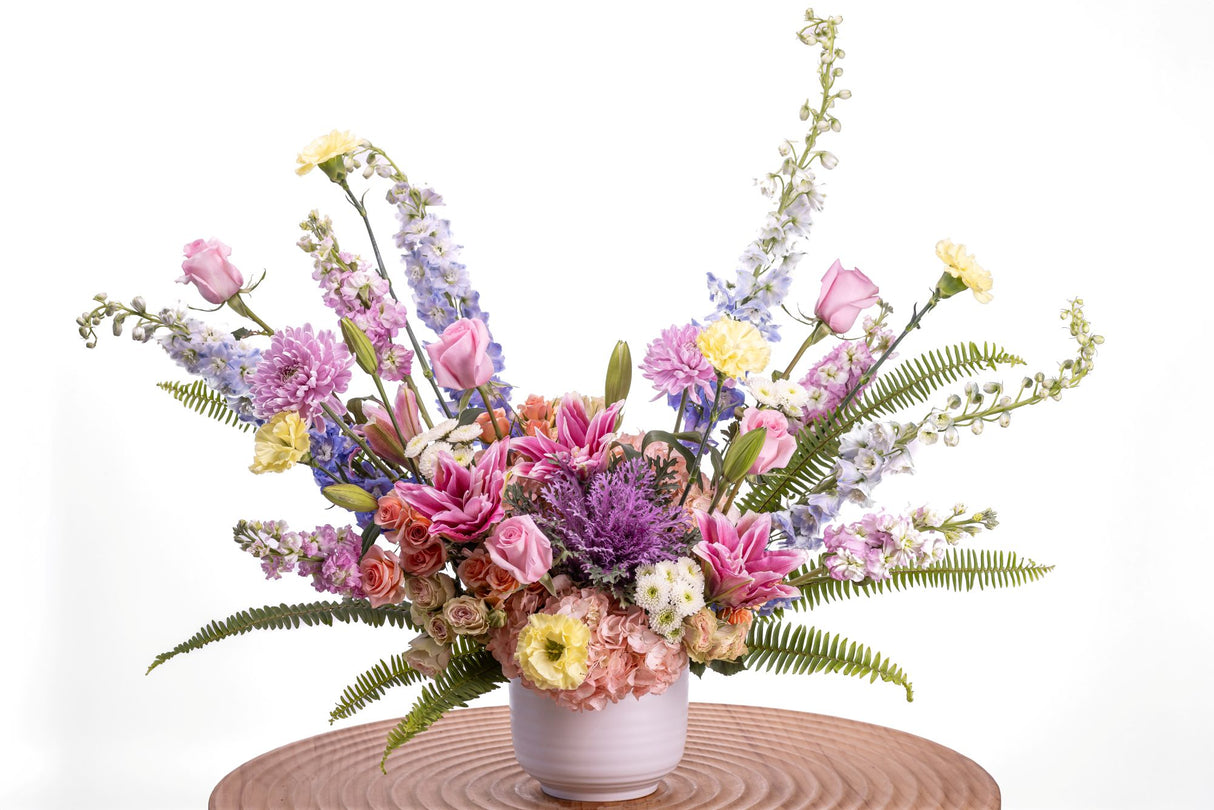 Serenade features pink roses, peach spray roses, pink lilies, tinted hydrangea, light blue delphinium, yellow carnations, lavender cushion poms, and white button poms arranged in a white ceramic vase with greenery.