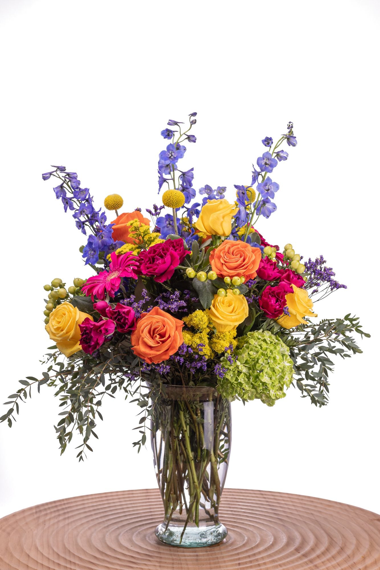 Showgirl features yellow and orange roses, hot pink spray roses and gerbera, blue delphinium, craspedia, limonium, solidago, green hydrangea, hypericum, and eucalyptus arranged in a tall Alessandra clear glass vase.