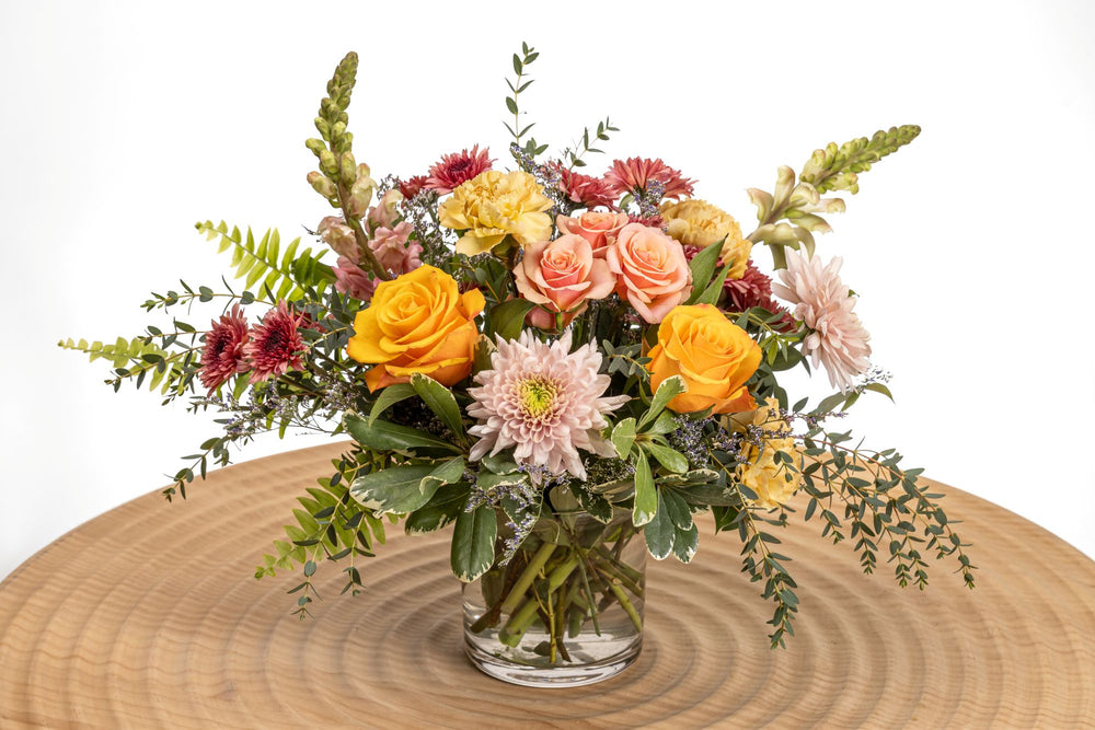Summer Sunset features pink tulips, Free Spirit roses, peach spray roses, peach carnations, pink larkspur, liatris, cremone, cushion poms, limonium, greenery and wispy sword fern in a glass cylinder.