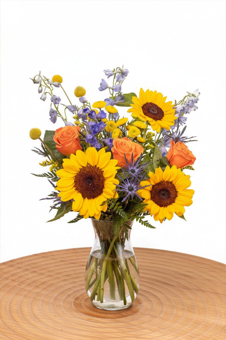 Sunbeam features sunflowers, orange roses, blue delphinium, craspedia, yellow poms, thistle, and solidago arranged with greenery in a glass vase.