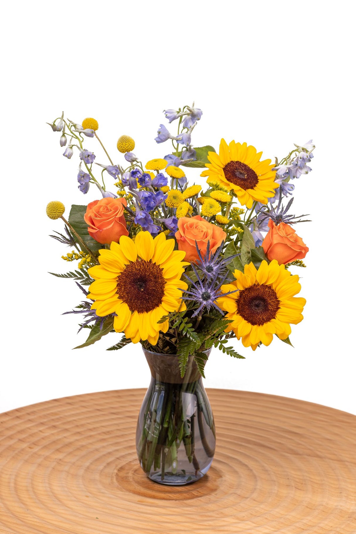 Sunbeam features sunflowers, orange roses, blue delphinium, craspedia, yellow poms, thistle, and solidago arranged with greenery in a glass vase.