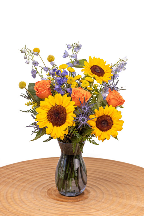 Sunbeam features sunflowers, orange roses, blue delphinium, craspedia, yellow poms, thistle, and solidago arranged with greenery in a glass vase.
