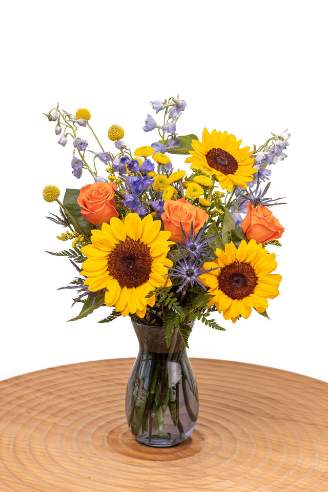 Sunbeam features sunflowers, orange roses, blue delphinium, craspedia, yellow poms, thistle, and solidago arranged with greenery in a glass vase.