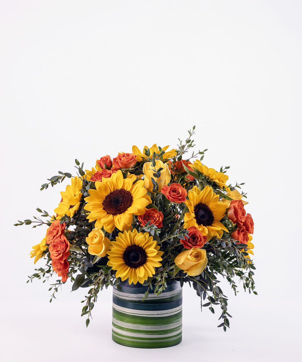 Bright floral arrangement with yellow roses, orange spray roses, and sunflowers in a clear glass vase
