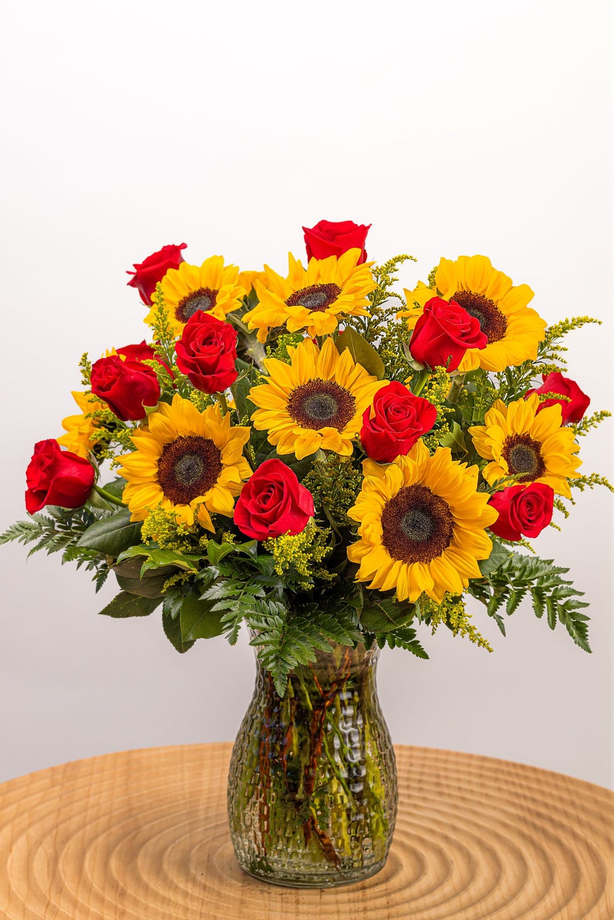 A cute flower arrangement featuring yellow sunflowers, red roses, and fresh greenery in a large, tall vase.