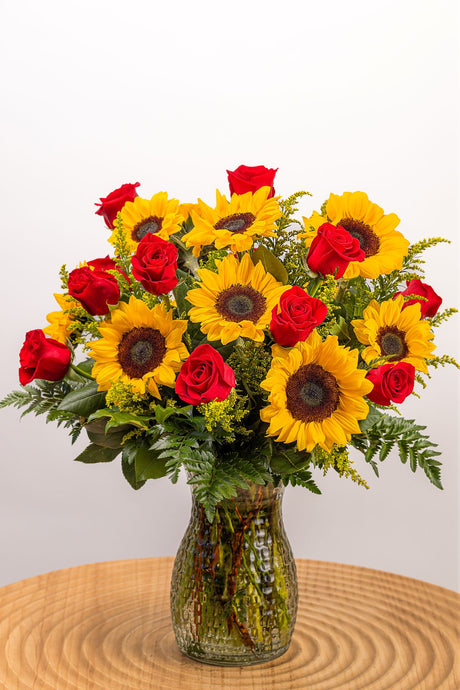 A cute flower arrangement featuring yellow sunflowers, red roses, and fresh greenery in a large, tall vase.