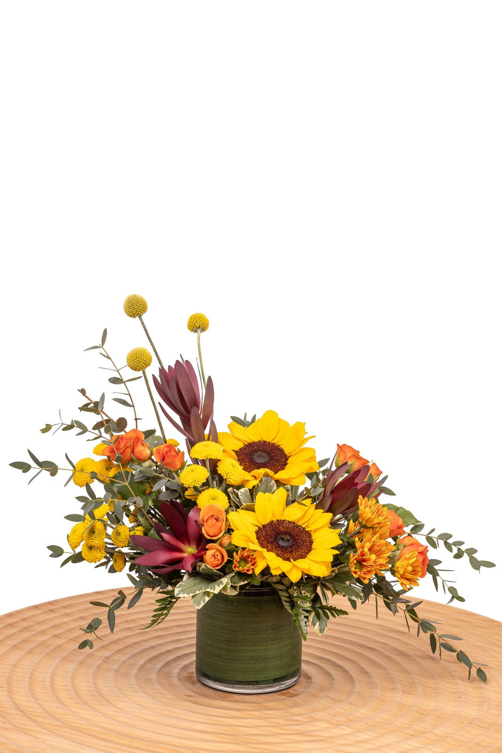 Topaz features sunflowers, orange spray roses, yellow craspedia, and safari sunset with lush greenery arranged in a glass cylinder vase.