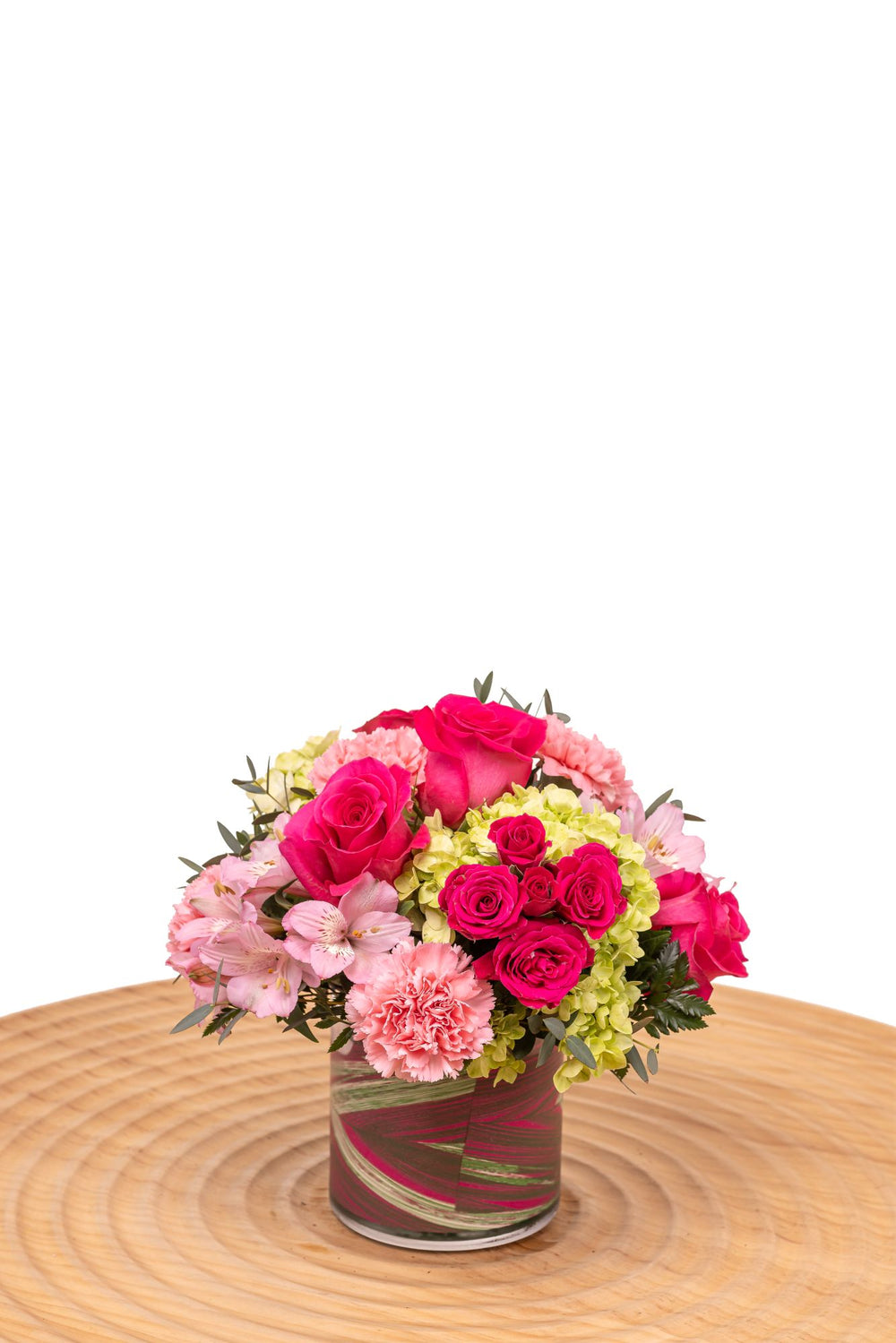 Tourmaline features hot pink roses, pink carnations, alstroemeria, and green hydrangea arranged in a glass cylinder vase wrapped with decorative leaves.